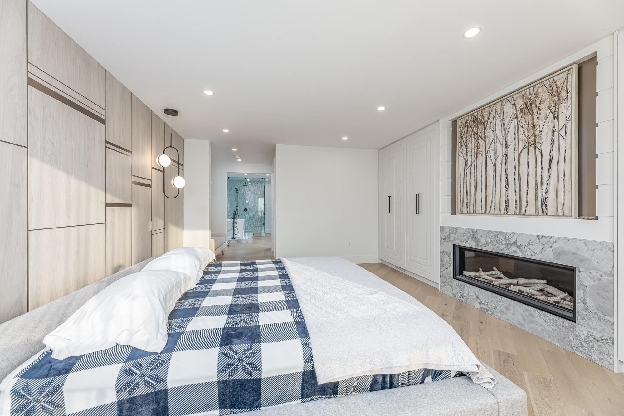 Primary bedroom in Ontario Airbnb rental — king bed, linear marble fireplace, custom wood panel wall, pendant lighting and ensuite bathroom beyond